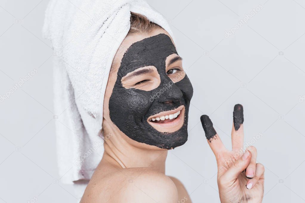 Horizontal studio portrait of happy young woman blink with eye with cosmetic black clay organic mask on her face, wears black white on hair. Female taking care of face skin, isolated on white wall