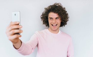 Beyaz stüdyo duvar arka plan karşı pembe elbise giymiş neşeli Kafkas erkek kıvırcık saçlı selfie mutlu gülümsemeyle için poz yatay portresi. Kişi, yaşam tarzı ve teknoloji kavramı.