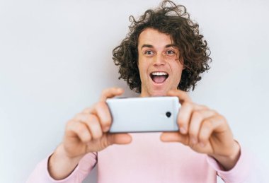 Yatay bir kadeh beyaz stüdyo duvar arka plan karşı pembe elbise giymiş neşeli Kafkas erkek poz için selfie mutlu gülümseme, kıvırcık saçlı. Kişi, yaşam tarzı ve teknoloji kavramı.