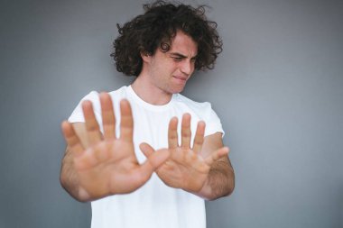 Reddi ve stop jest, yakışıklı kıvırcık saçlı erkek portresi gösterir beyaz t-shirt, pozlar gri beton stüdyo duvar arka plan karşı giyen kızgın ve mutsuz ifade vardır. Metin için yer kopyalayın