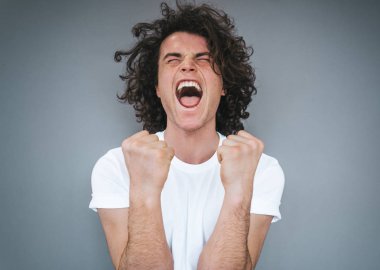 Heyecanlı başarılı yakışıklı genç erkek beyaz t-shirt giyiyor, clenches yumruk sevinç, çığlık açılır ağız ile yaygın olarak gri studio arka plan üzerinde izole, mutluluk ile söyler gibi. Reklam alanı