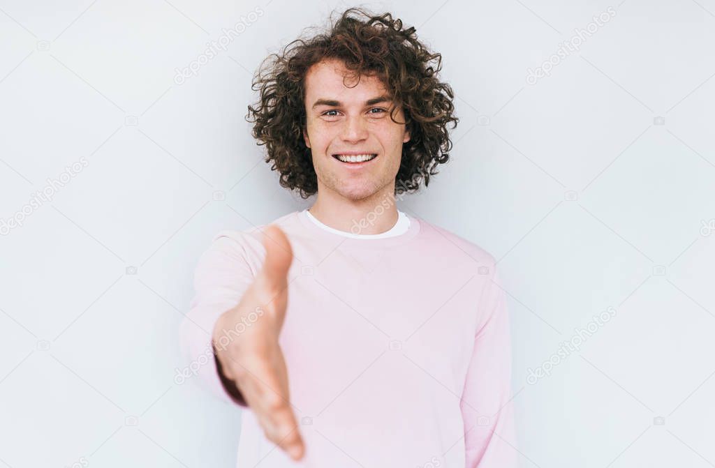 Portrait of young cheerful man with trendy curly hairdo smiling broadly while stretching hand at camera in handshake gesture, standing over white background. People and body language concept