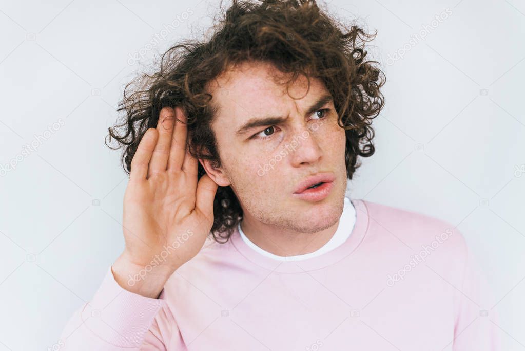 Closeup shot of handsome eavesdropping male with curly hair placing hand on ear asking someone to speak up, isolated over white background. Stylish man which overhears conversation. Copy space