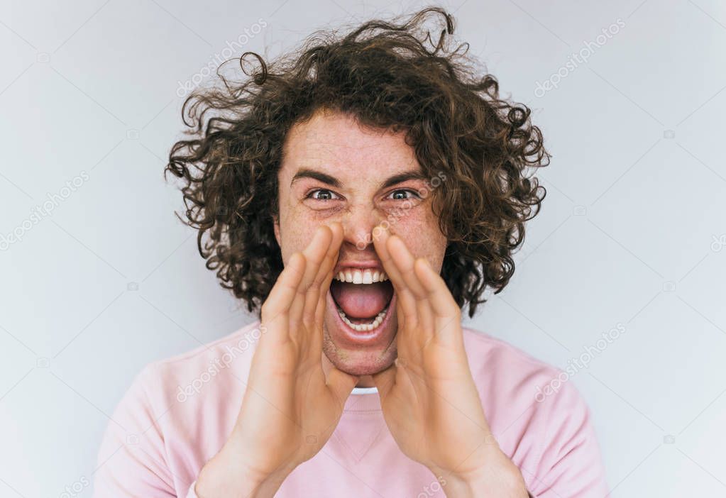Horizontal shot of handsome angry male with curly hair opens mouth widely, screams in panic with hands on mouth, being in stressful situation posing on white studio wall. People and emotion concept.