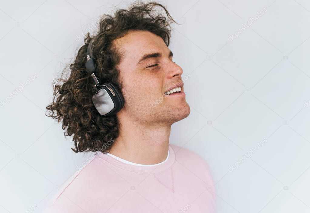 Side view portrait of handsome smiling young male model with curly hair and freckles wearing pink clothes, listening music on headphones, isolated on white studio wall. Copy space for advertisement