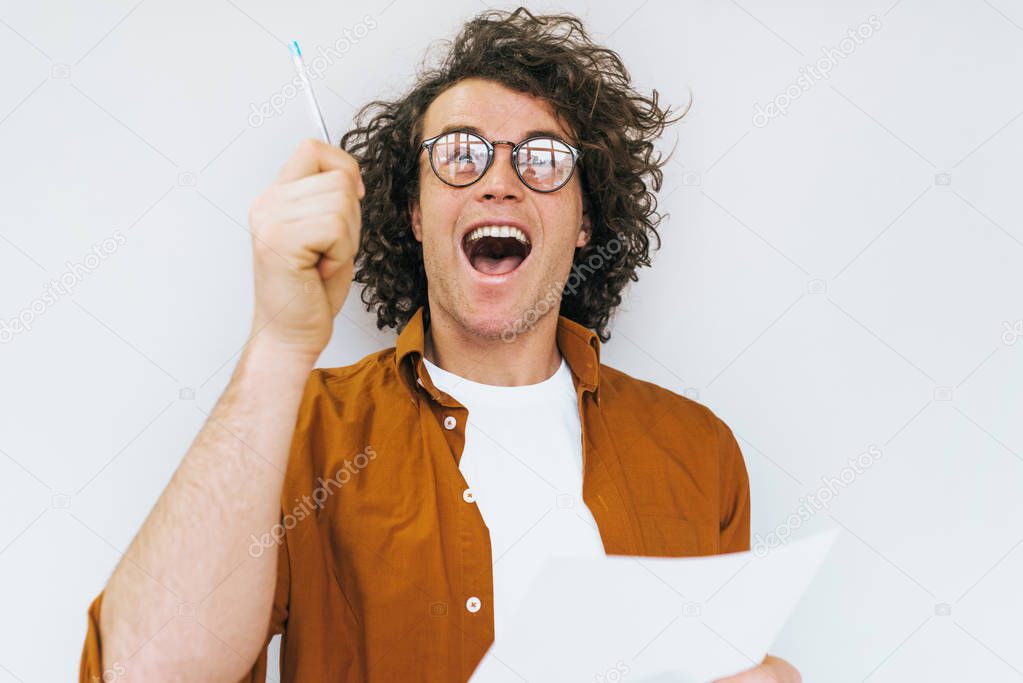 Portrait of happy cheerful student with curly hair, writes on paper new checklist for exams posing over white studio wall. Smiling succesful worker man wears round spectacles makes notes in notebook 
