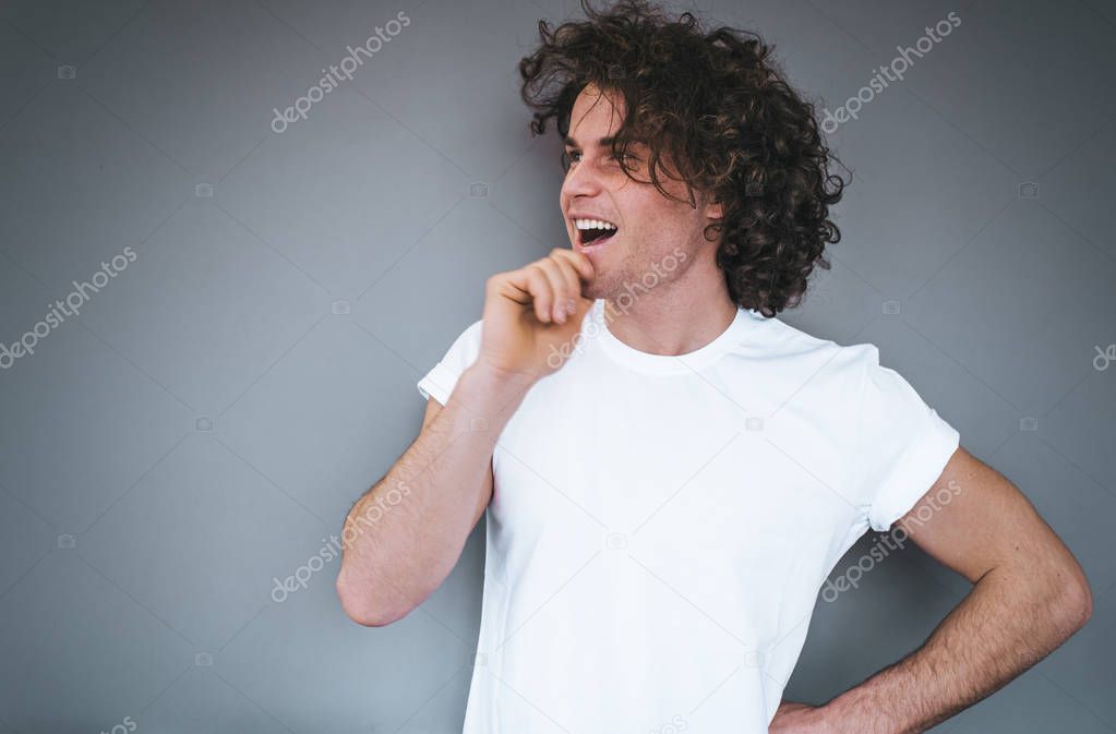 Portrait of handsome happy thinking young male with curly hair, wearing white t-shirt, looking away posing against grey background. Copy space for advertisement. People and emotion concept.