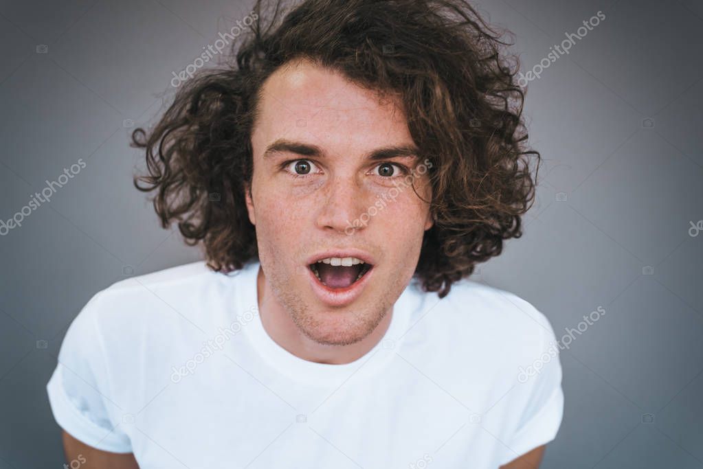 Closeup horizontal portrait of curly hair amazed handsome man has excited expression, recieve or reading good news. Happy pleased male wears white t-shirt isolated on grey studio wall. Copy space