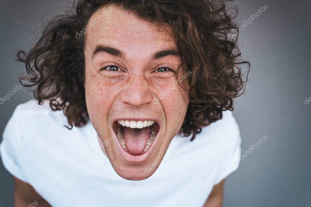 Closeup portrait of attractive curly hair amazed handsome man has excited expression with open mouth screams loudly. Happy pleased male wears white t-shirt isolated on grey studio wall. Copy space