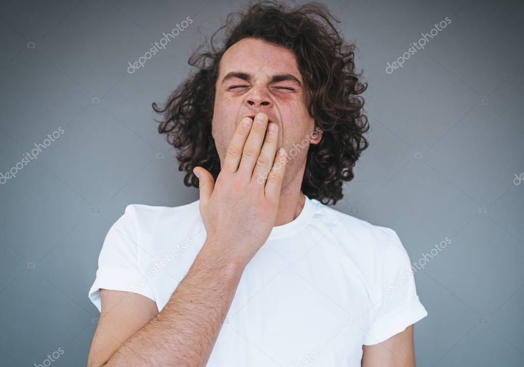 Portrait of tired or bored Caucasian young male model with curly hair covering mouth while yawning, feeling exhausted after overwork. Student stylish man having sleepy look after lessons.