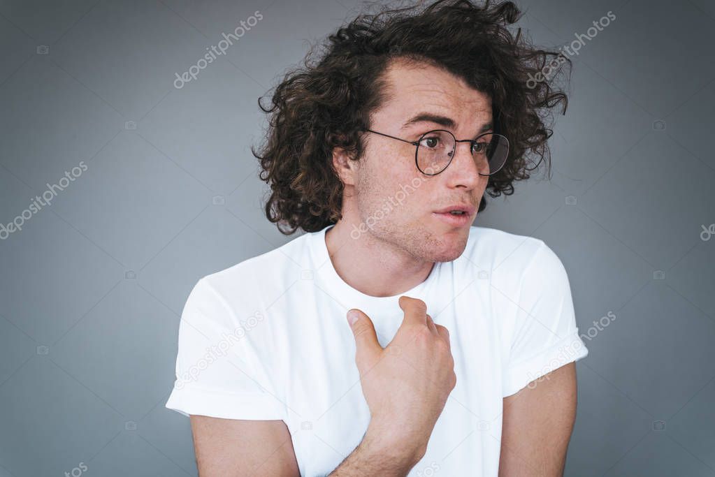 Confused handsome young man with curly hair points at himself being uncertain to be chosen, wears white t-shirt and spectacles. Amazed male looking away indicate at himself with hand on grey wall
