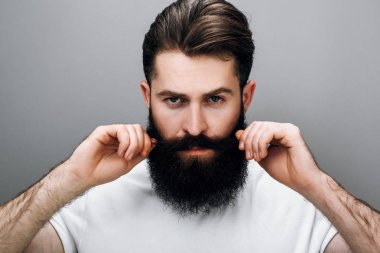 Horizontal close up portrait of brutal handsome young bearded male corrects his moustache and beard, looking at the camera, wears white tshirt, on a gray studio wall. Young barber with trendy beard. clipart