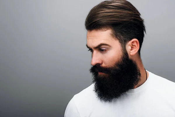 Side view of handsome brutal masculine young bearded male looking aside, posing on a gray studio background. Portrait of young European fashion barber with trendy beard.