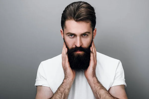 Yakışıklı erkek genç sakallı adam yatay portre sakal ve gri studio arka plan üzerinde kamera bakıyor yandan tutuyor. Trendy sakallı genç Avrupa hipster portresi.