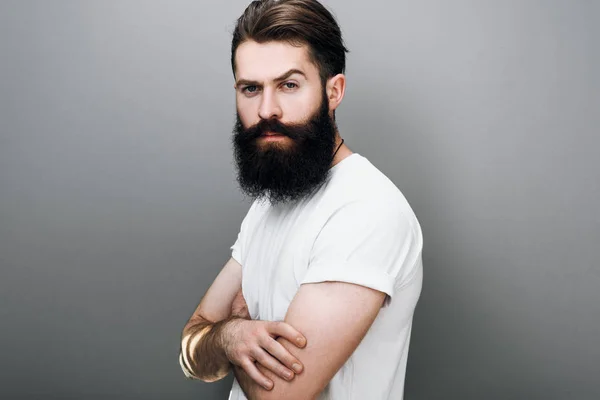 Kalın sakal ve bıyık, acımasız Kafkas Erkek kılığında casual beyaz t-shirt gri arka plan üzerinde poz içinde. Onun yüzünde kendinden emin ifade ile Avrupa man modeli sakallı
