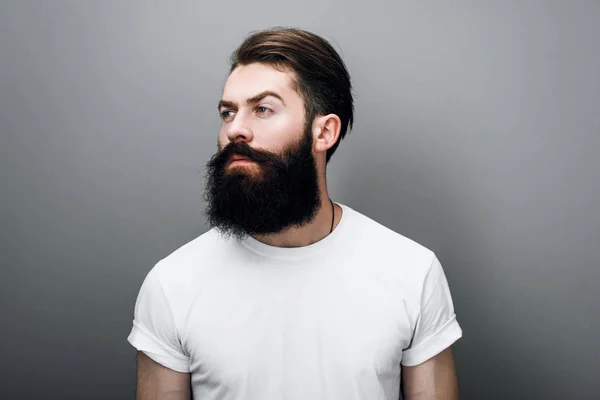 Brutal hipster Caucasian male with thick beard and mustache, dressed in casual white t-shirt posing on gray background. Bearded European man model with confident expression on his face