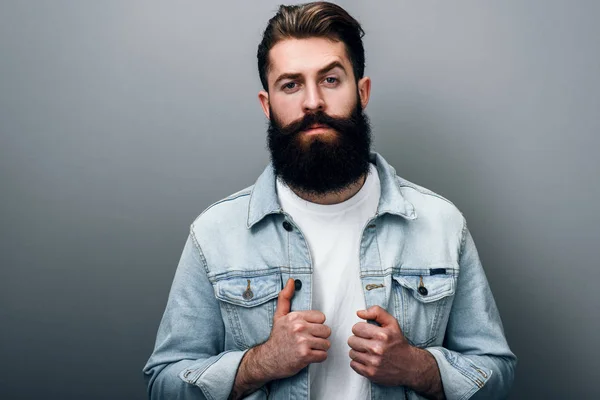 Fotoğraf makinesi ciddi ve neşeli bir görünüm ile bakan yakışıklı şık genç Avrupa erkek trendy denim ceket kalın sakallı. Kuaför adam gri studio duvara poz.
