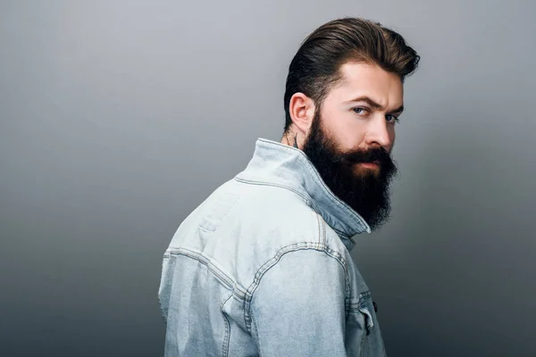 Dikiz kamera ciddi bakışla bakıp şık acımasız genç Kafkas erkek moda kot ceket kalın sakallı. Yakışıklı Kuaför adam gri studio duvara poz