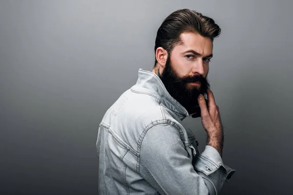 Arka görünüm portre fotoğraf makinesi ciddi göz ile bakan şık acımasız genç Kafkas erkek moda kot ceket kalın sakallı. Yakışıklı Kuaför adam gri studio duvara poz