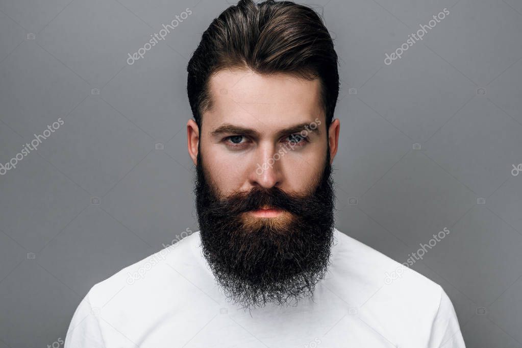 Horizontal close up portrait of handsome brutal masculine young bearded male looking at the camera, posing on a gray studio background. Portrait of young European fashion barber with trendy beard.