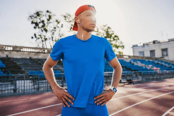 Portrait of sporty professional male athlete standing with his hands on waist looking aside on sunlight. Caucasian sprinter stands in line at the athletics track in