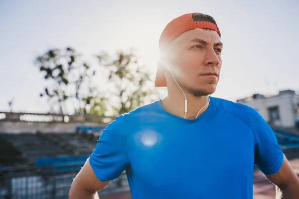 Close up portrait of sporty professional male athlete standing with his hands on waist looking aside on sunlight background. Caucasian sprinter stands in line at the track in stadium. Sport, people