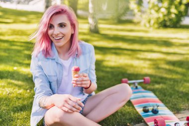 Çok Avrupa güzel gülümseyen kadın dondurma elinde yeşil çim arka plan mavi gömlekli Park'ta oturuyor ile pembe saç üfleme. Sportif erkek longboarding sonra rahatlatıcı.
