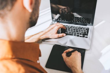 Kalem ve tablet masasında genel laptop çalışma, yazarak, çizim, ekrana bakarak genç işadamı yakından görmek arka. Defter, onun işyerinde beyaz üzerinde oturan kullanarak erkek serbest meslek.