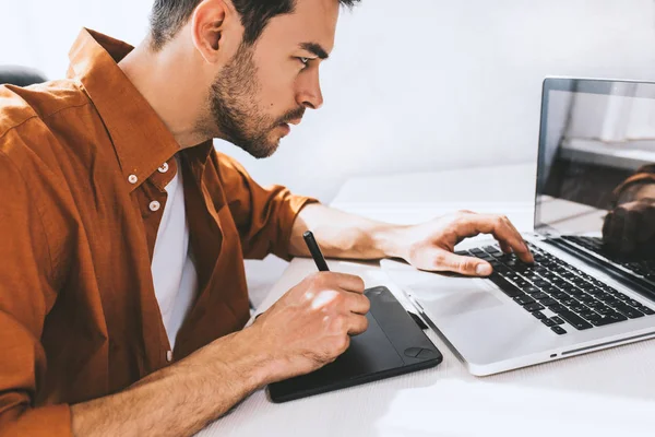 Ciddi ve düşünceli ifade ile belgili tanımlık laptop önünde oturan erkek serbest meslek yoğunlaşmıştır. Genç tasarımcı ya da ofiste oturan iş için kalem ve bir dizüstü bilgisayar kullanarak gömlekli mimar.