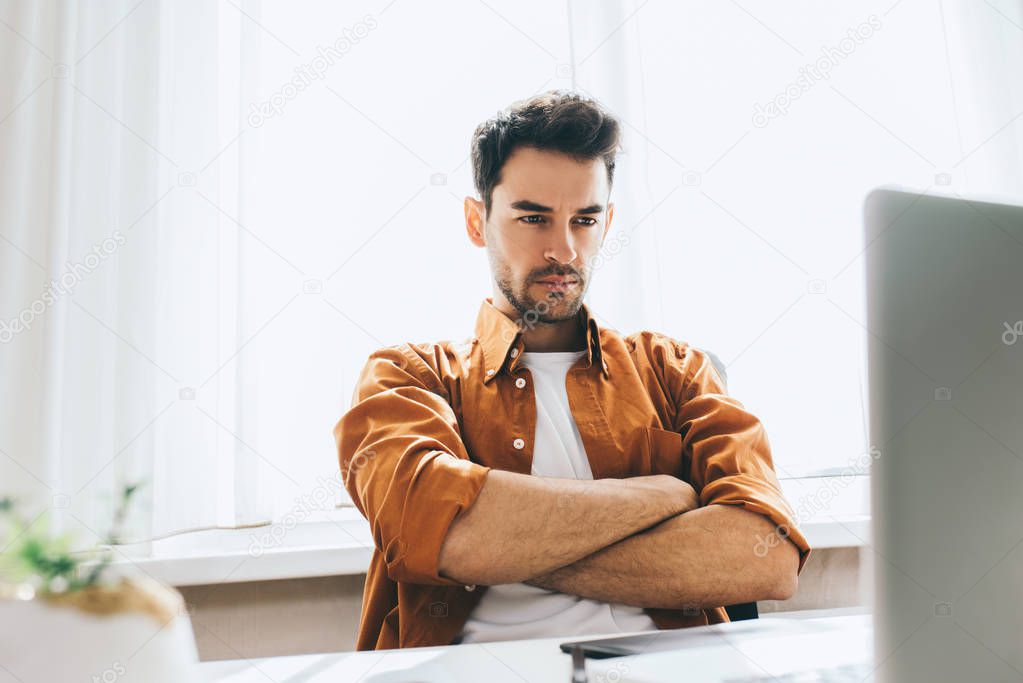 Serious and handsome male freelancer sitting in front of the computer with serious and thoughtful expression crossed his arms. Businessman think about resolves some problems in the office.