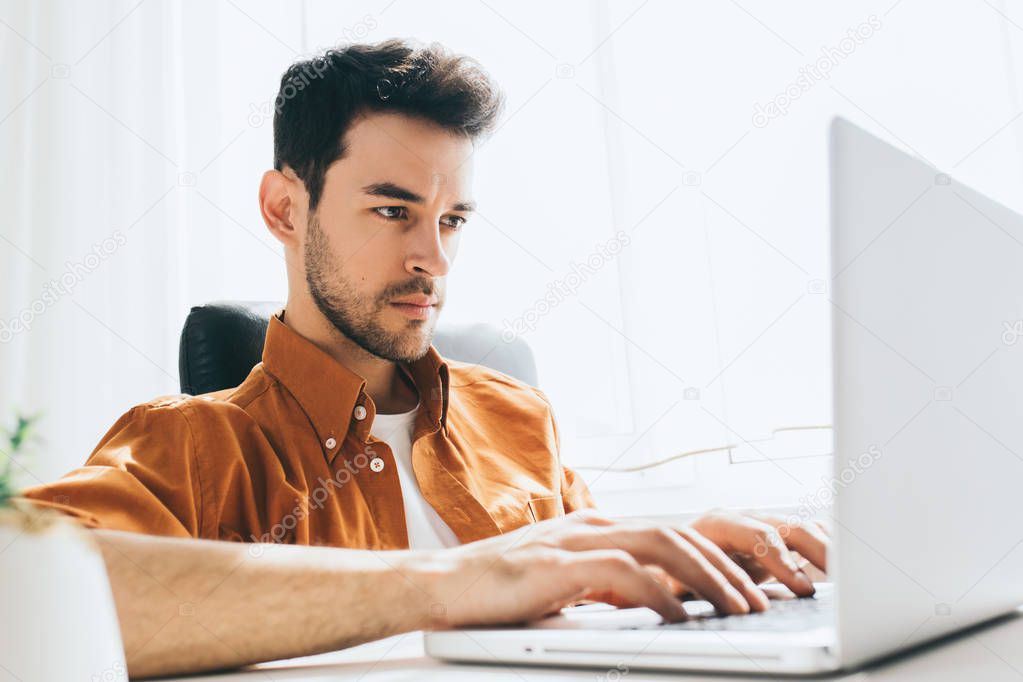 Attractive and confident male freelancer sitting in front of the computer with serious and thoughtful expression. Young businessman in shirt typing on laptop pc sitting in the office.