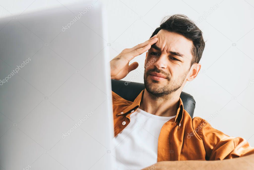 Young Caucasian man in casual clothes have headache behind computer. Frustrated young handsome man worried about problems in business. Stressed businessman after long web conversation.