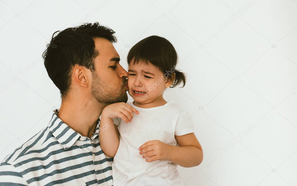 Handsome father holding and kiss his crying daughter. Young dad embrace his sad child. Attractive man support his unhappy toddler girl. Childhood and fatherhood relationship. Lifestyle family.