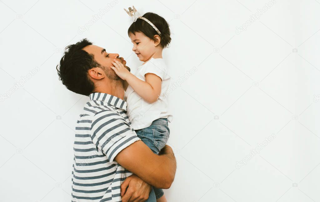 Funny dad and pretty daughter play and cuddle together against white background. Happy sincere relationship of parent and child. Family moments of father and toddler girl's birthday. Parenthood care.