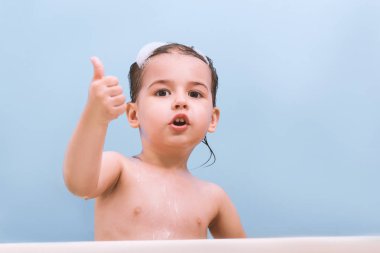 Neşeli mutlu bebek, yürümeye başlayan çocuk bir banyo baş parmakları. Köpük ile bir küvet içinde küçük bir çocuk. Mavi arka plan banyo çocukta gülümseyerek. Bebek yıkama ve banyo. Hijyen ve sağlık.