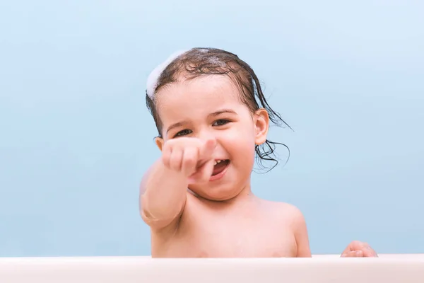 風呂に入って泡で遊んで楽しい陽気な幸せな幼児赤ちゃん バスタブで 幼い子供は 彼の指でポイントします 青の背景に浴室で子供を笑っています 幼児が洗濯と入浴 健康管理 — ストック写真