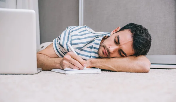 Young Caucasian freelancer business man sleeping near to laptop with eyes closed while lying on the floor home and notice paper. People, lifestyle, technology, business and communication concept.
