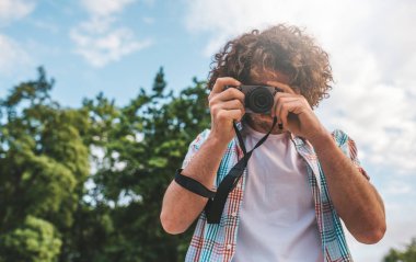 Genç Turizm erkek ile dijital fotoğraf makinesi çekim, bir doğa ve gökyüzü arka plan üzerinde. Yakışıklı adam kıvırcık saçlı doğa tatil gezisinde keşfetmek. Seyahat, insanlar ve yaşam tarzı kavramı 