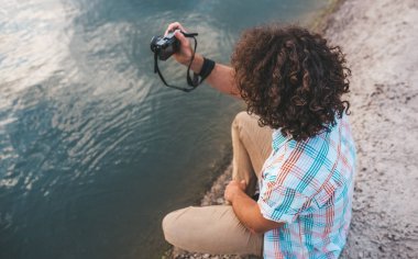 Fotoğraflar Doğa onun dijital fotoğraf makinesi üzerinde denetimini kıvırcık saçlı Genç erkek yan görünüm. Yakışıklı adam gölün yanında poz dijital fotoğraf makinesi ile rahat gömlek giyer. İnsanlar, seyahat, yaşam biçimi