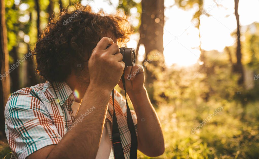 Horizontal side view of handsome male with curly hair taking photos of nature on his digital camera. Young traveler man with digital camera posing in the forest on sunset. People, travel, lifestyle