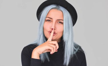 Portrait of blue hair female wears black clothes, shows silence sign, asks to keep information confidential, has pretty expression, isolated on grey studio background. Secret gesture, shhh.  clipart