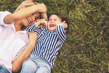 Genel gider görüntü oynama ve sevimli küçük kızı ile açık yeşil çim üzerinde yalan mutlu güzel sarışın kadın. Sevgi dolu anne ve kızı parkta beraber vakit. Anne ve çocuk dışarıda eğlenmek