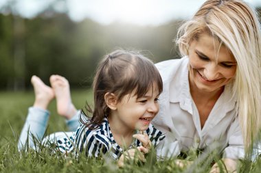 Yeşil çim üzerinde yalan ve parkta güzel annesi ile oynarken mutlu küçük kız görüntüsü. Kadın ve küçük kızı tatil keyfini çıkarın. Anne ve çocuk açık havada eğlence vardır. Anneler günü