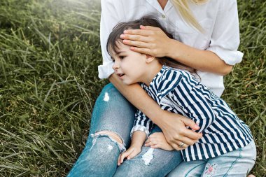 Sevimli küçük kız açık havada annesinin ayak üzerinde yalan görüntüsü. Sevgi dolu anne ve kızı parkta yeşil çimenlerin üzerinde oturuyor. Anne ve çocuk sarılma ve eğlenceli vardır. İyi bir ilişki. Anneler günü