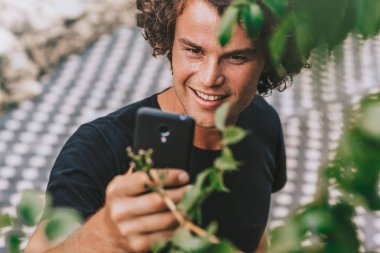 Gülümseyen adam açık havada duran closeup çekim üstünde hareket eden telefon görüşme. Cep telefonundan beton gri arka plan üzerinde wirless kullanarak sokaktan dışarı dinlenme kıvırcık saçlı Genç erkek