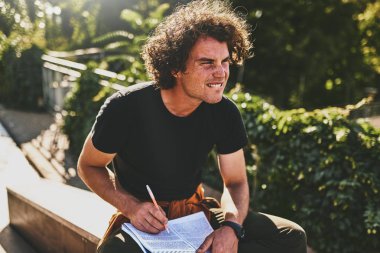 Kıvırcık saçlı yakışıklı öğrenci adam kağıt ve sınavları için hazırlık bazı bildirimler yazma sokak şehir oturan siyah t-shirt giymiş. Serbest meslek sahibi işadamı yeni projeler için planlar yapar.