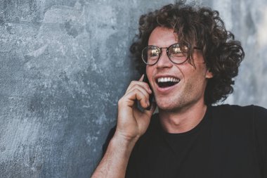 gülüyor, kıvırcık saçlı cep telefonu gri duvarın karşısında dururken konuşurken mutlu yakışıklı adam