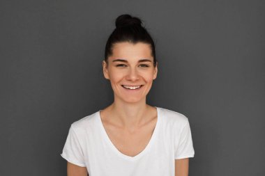 Mutlu bir şekilde bazı olumlu haberler alınırken gülümseyen düğüm saçlarını giyiyor neşeli muhteşem genç esmer bir kadın portresi. Güzel kız kameraya neşeli gülümseme ile seyir beyaz t-shirt giymiş