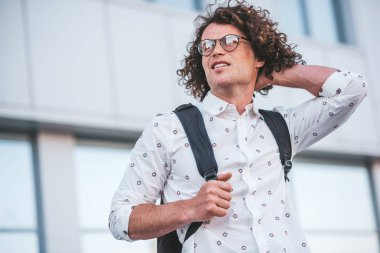 Alt görünümü atış yakışıklı genç erkek öğrencinin nereye gideceğini düşünerek sokakta arka plan binasında duran sırt çantası. İş adamı rahat beyaz gömlek ve gözlük takıyor. İnsanlar