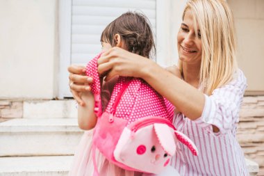 Çocuk kızına anaokuluna açık oluyor üzerinde sırt çantası koymak sırt çantası hazırlama mutlu gülümseyen anne. Sevimli küçük kız anne ev karşı ilk okul öncesi gün önce ile arka görünümü